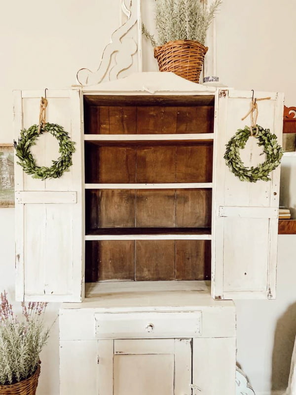 Empty white antique cabinet in bedroom of farmhouse with boxwood wreaths on doors.