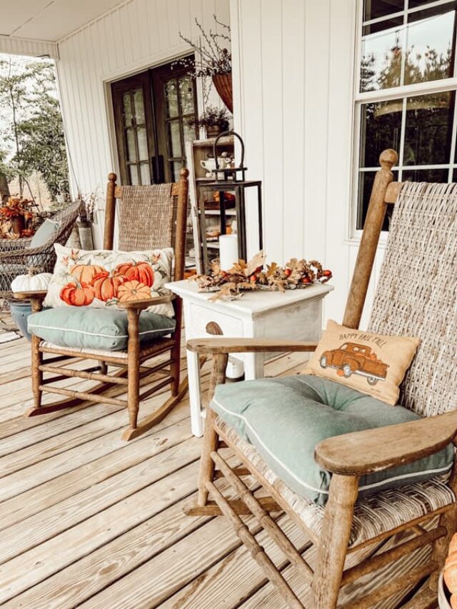 two rocking chairs on front porch with fall pillows and blue cushions.  White center table with lantern and fall arrangement.