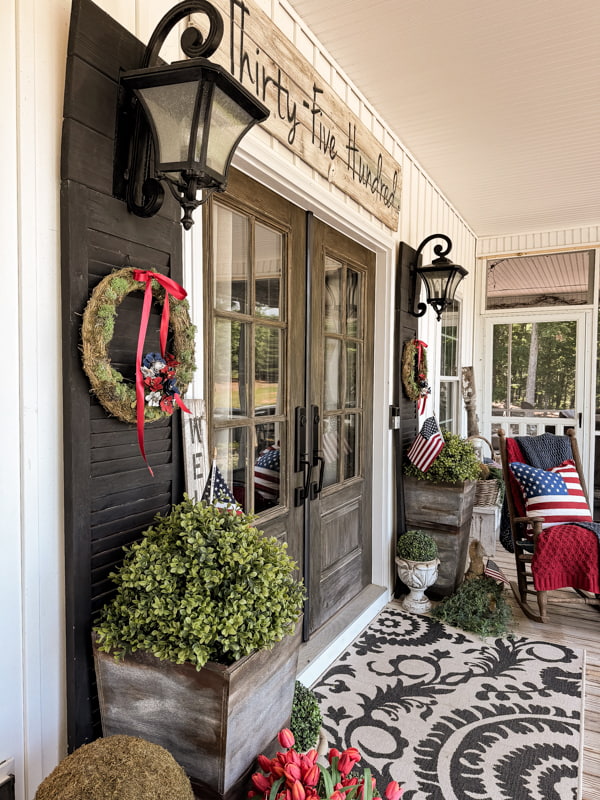 DIY moss wreaths with pinecone red, white and blue flowers beside front doors with planters filled with faux boxwood.