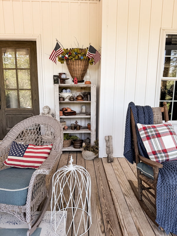 Rustic primitive cabinets with a wall basket filled with greenery and flags on front porch wth rockers in front.