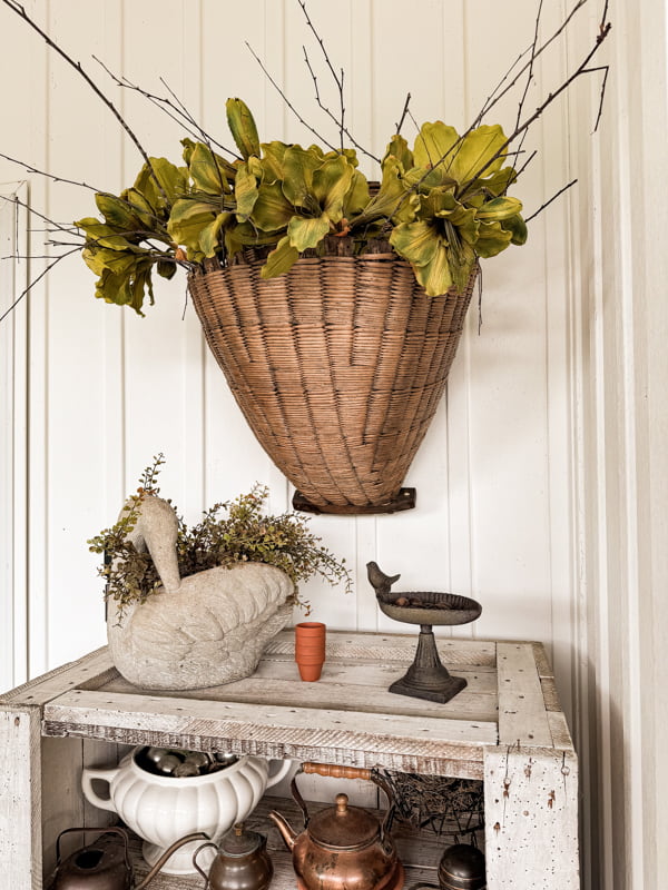 Hanging wall basket on porch with green flowers and gathered stems.  Concrete swan planter with greenery.