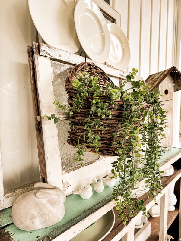 thrift store wall basket with greenery hanging on old window with vintage ironstone plates and DIY birdhouse.  