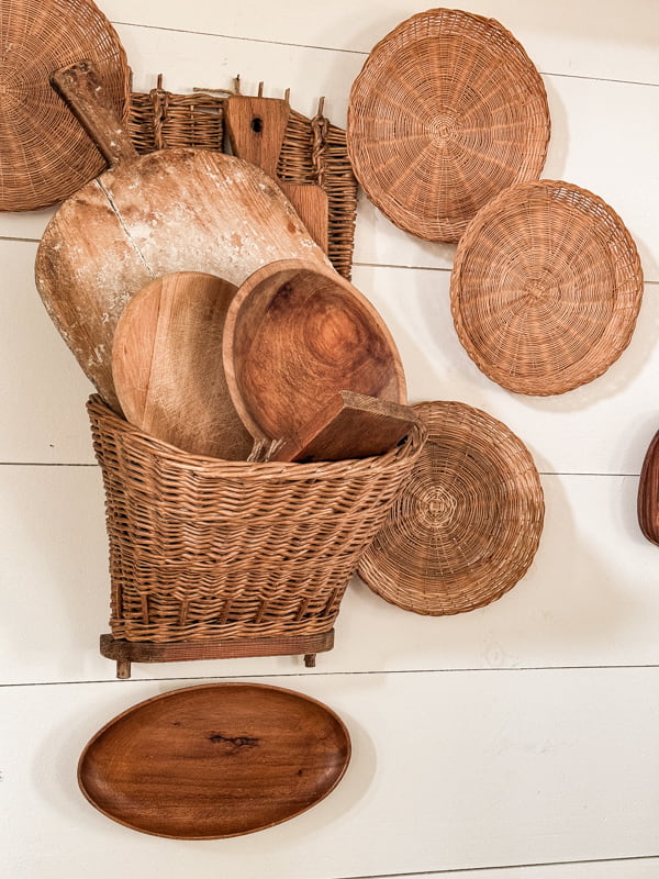 Grape hod gathering basket filled with breadboards and wooden bowls on dining room wall with wicker plate trays 