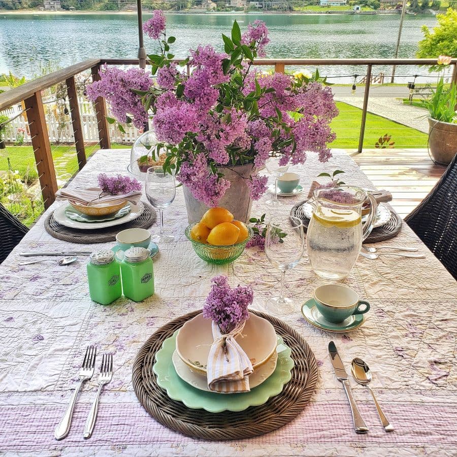 Outdoor living space deck overlooking water with table set with lilacs and vintage dishes.  