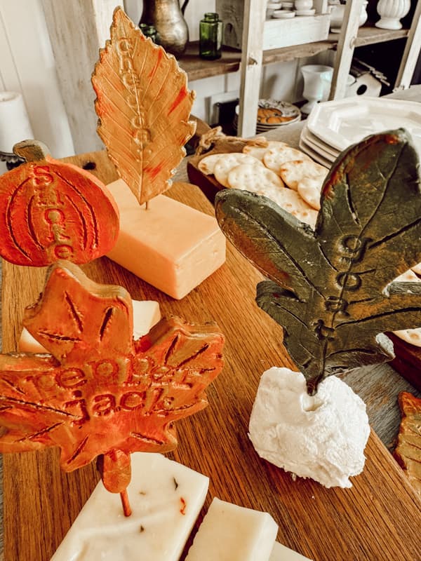 Handmade cheese markers in the shape of fall leaves and pumpkins on cheese board with crackers