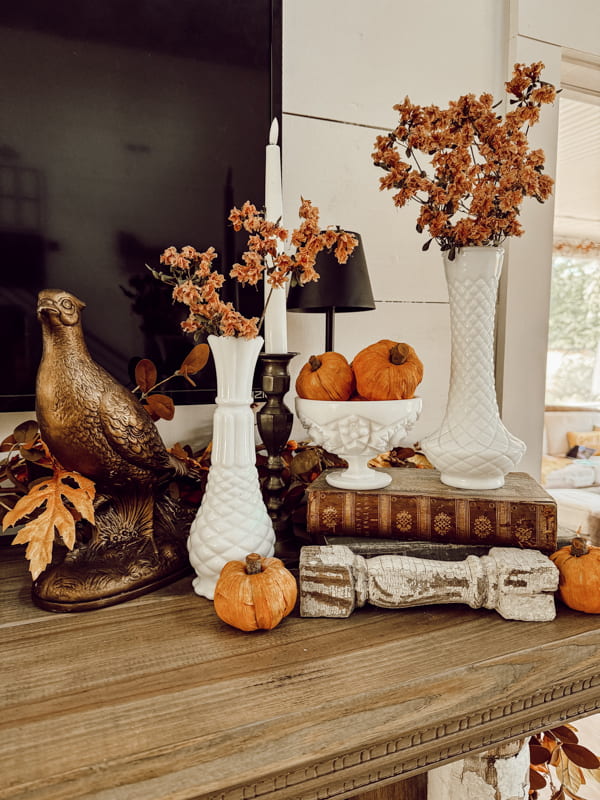 White milk glass bud vase for fall mantel decorations.  a pheasant and orange pumpkins with old books.