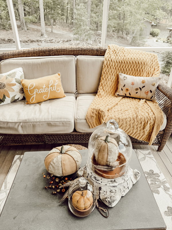 Coffee table with fabric pumpkins under glass cloches with outdoor sofa filled with cozy fall blankets and throw pillows.  