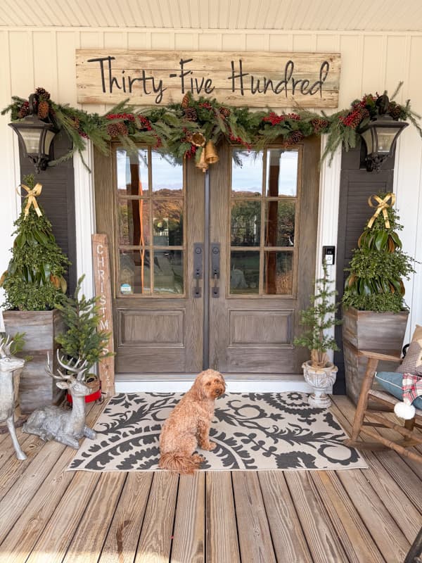 Rudy on front porch of farmhouse decorated for Christmas with DIY Greenery Trees and fun garland above the doors.  