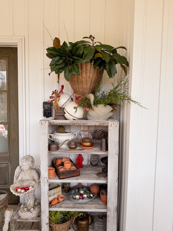 Primitive outdoor cabinet filled with thrift store finds and a wall hanging basket filled with foraged magnolia leaves. 